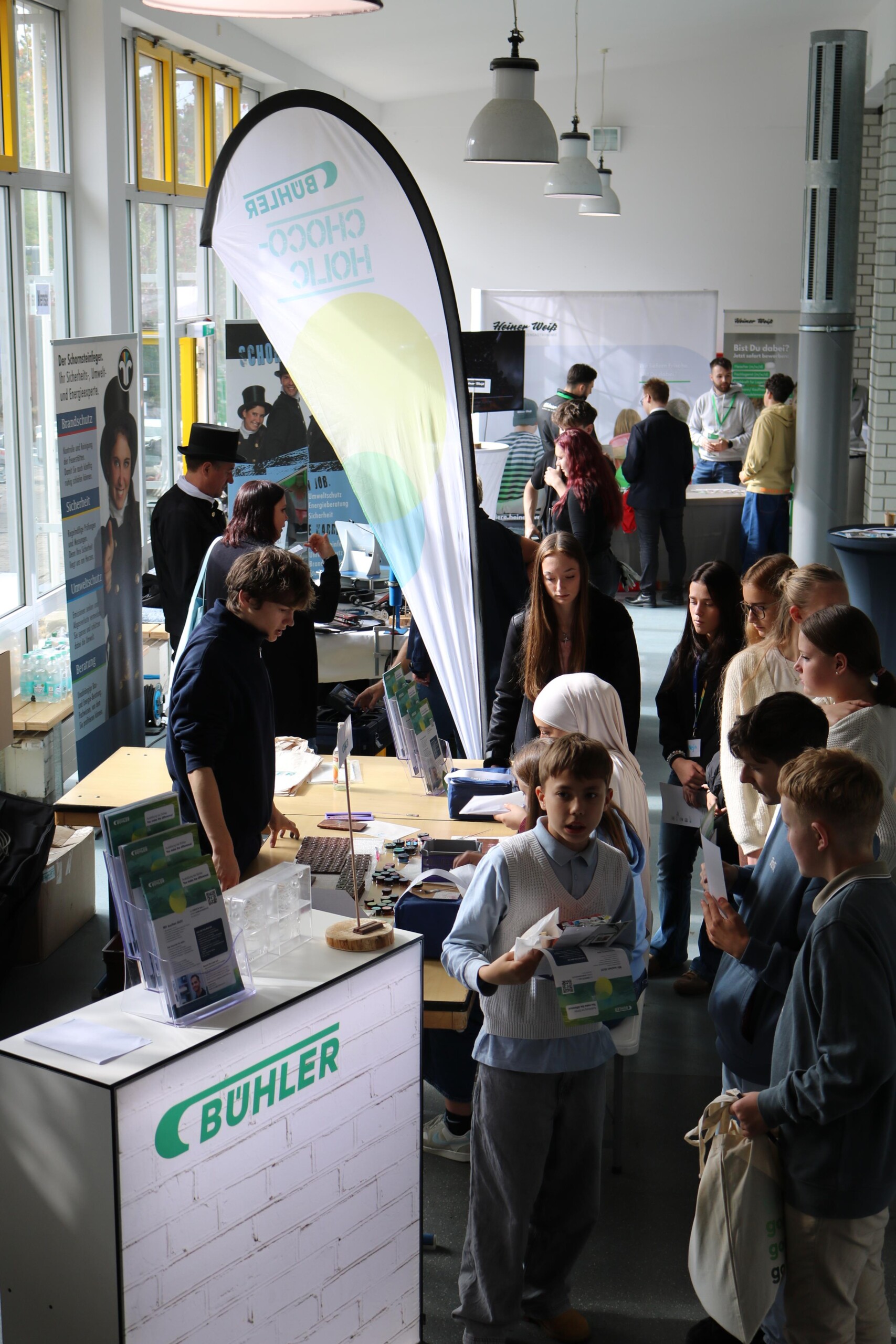An einem Messestand mit einem großen „Bühler“-Logo stehen Jugendliche und Erwachsene dicht gedrängt beisammen. Sie informieren sich, sprechen miteinander und betrachten Materialien auf den Tischen.