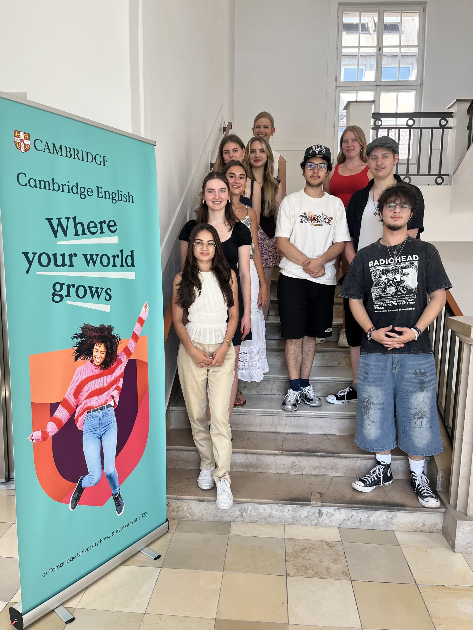 Die Teilnehmer:innen des Cambridge-Projektkurses stehen gemeinsam mit Lisa Dornseifer im Englischen Institut in Köln an einem Cambridge-Banner auf einer Treppe.