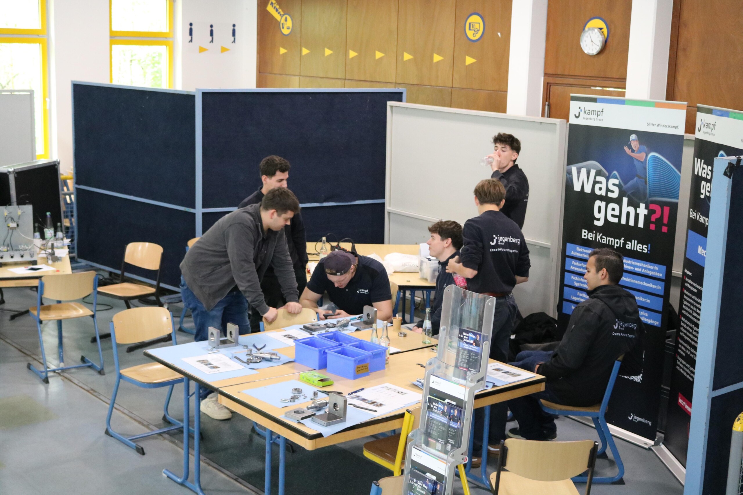 Mehrere junge Männer sitzen und stehen an einem Tisch, auf dem technische Bauteile liegen. Sie bereiten Präsentationen vor und sprechen miteinander.