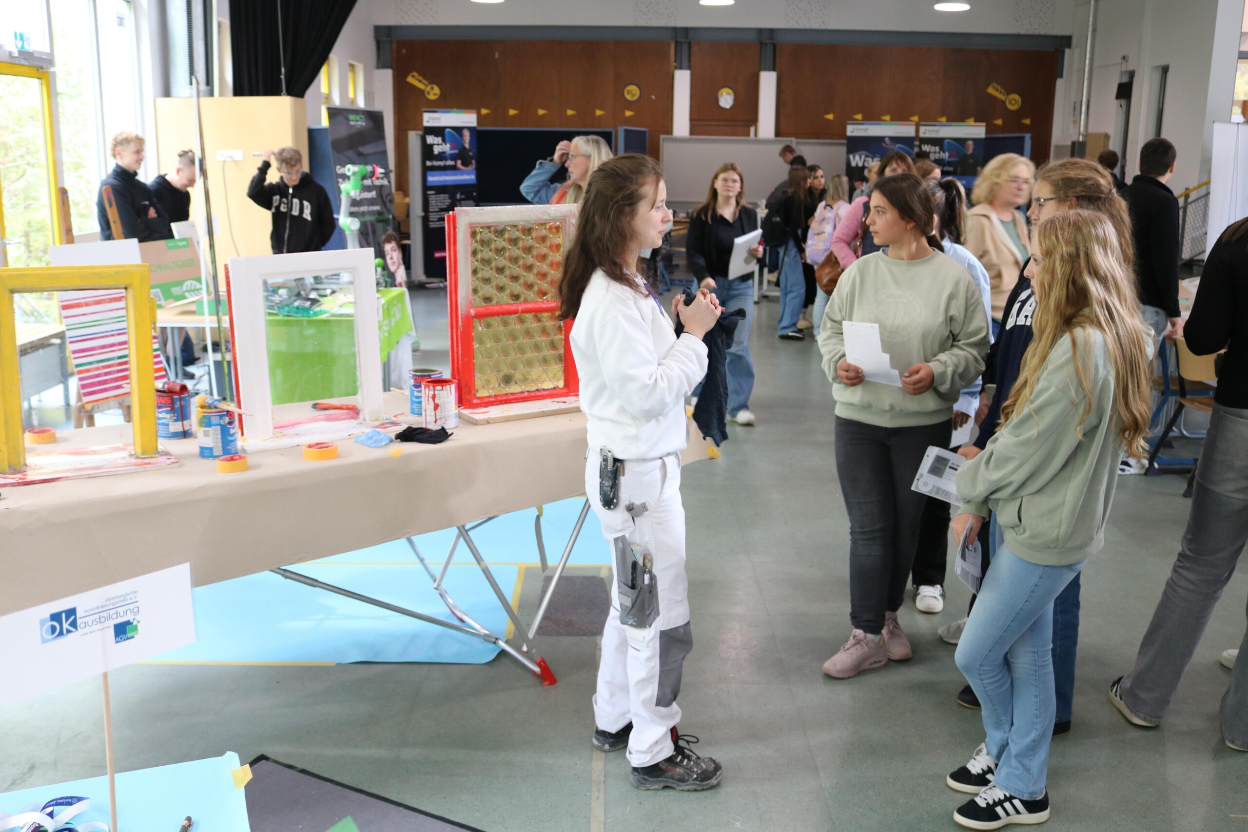 Eine junge Frau im weißen Handwerkeranzug erklärt einer Gruppe von Schülerinnen etwas an einem Stand mit bunt gestrichenen Fenstern.