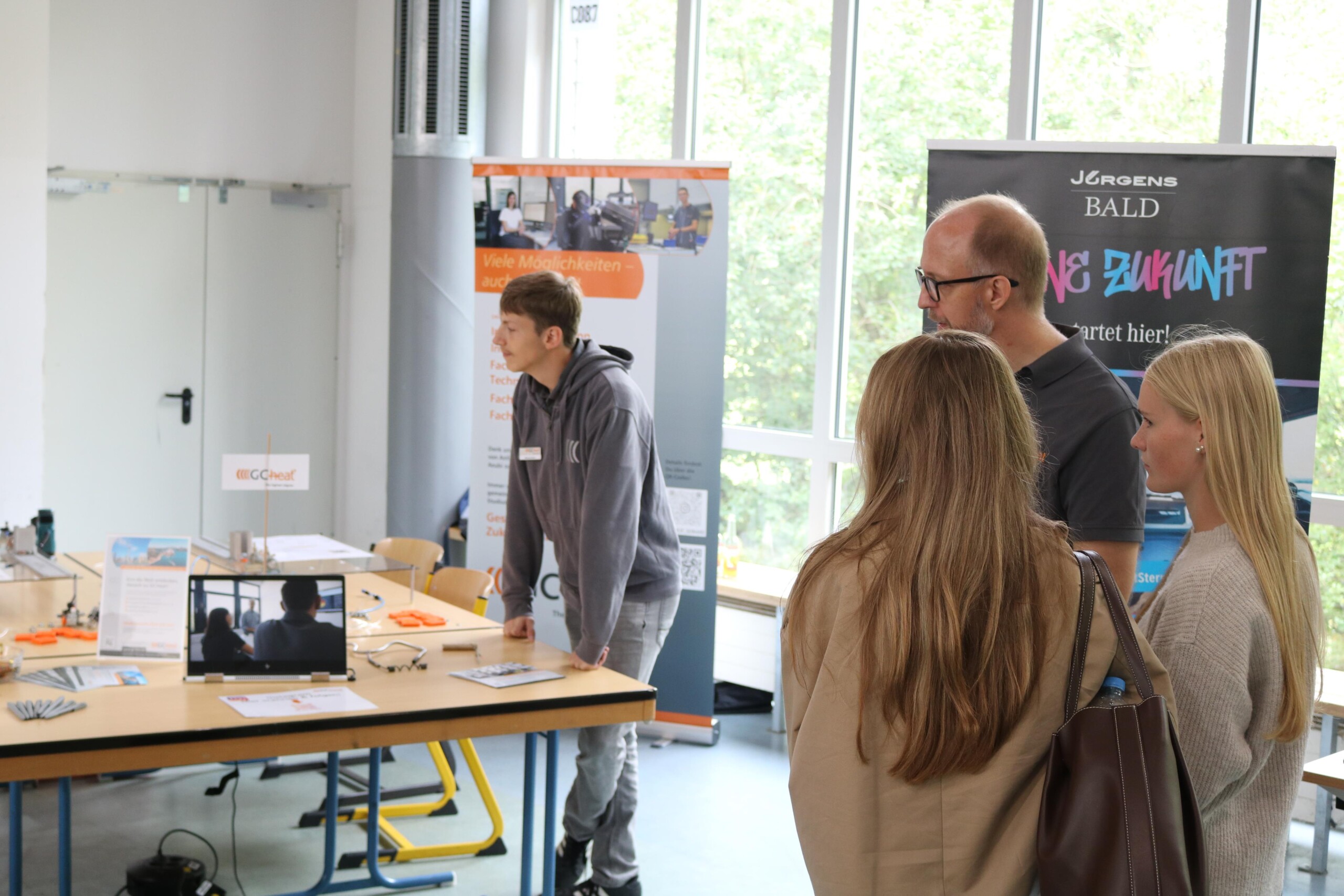 Drei Personen hören einem jungen Mann an einem Stand zu, auf dessen Tisch ein Laptop und Infomaterialien stehen.