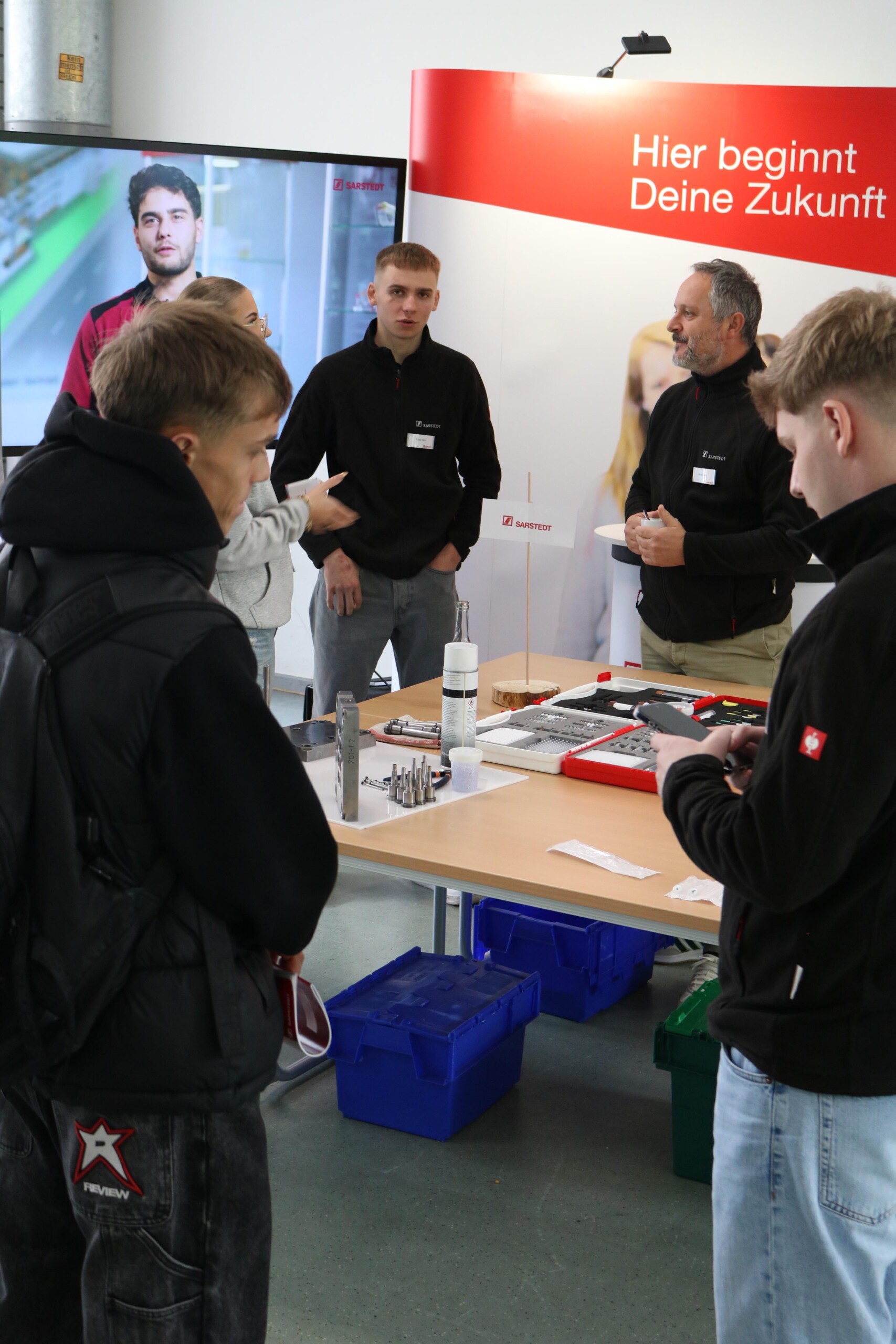 Jugendliche stehen vor einem Stand mit Werkzeugen und technischen Bauteilen. Zwei Aussteller im schwarzen Pullover beantworten Fragen.