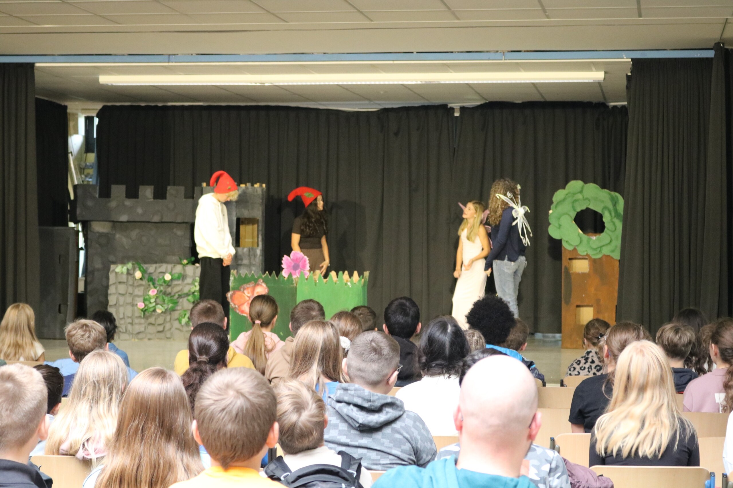 Der Kurs „Darstellen und Gestalten“ der Jahrgangsstufe 9 spielt eine Theaterszene auf der Probebühne im Forum. Im Bühnenbild sind eine Burgmauer, Blumen und ein Baum zu sehen. Zwei Schüler tragen rote Zipfelmützen, eine Schülerin spielt eine Fee, eine weitere einen Fabelwesen-Charakter. Vor der Bühne sitzt das junge Publikum der 5. Klassen und verfolgt die Aufführung aufmerksam.