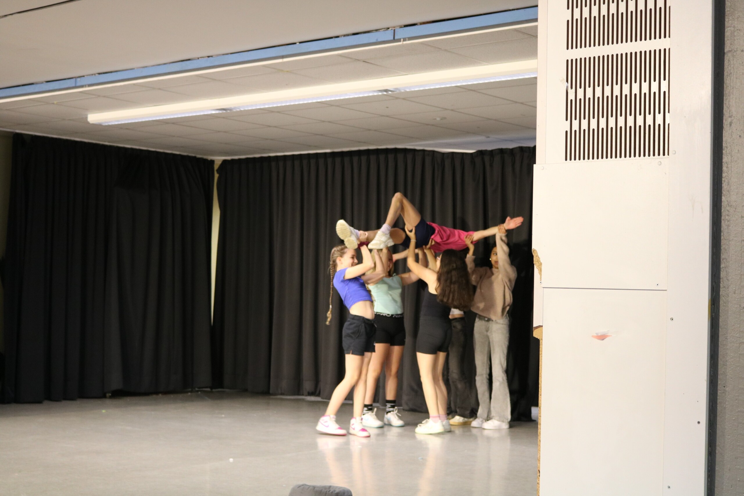 Schülerinnen der Klasse 6b präsentieren ihre Tanzakrobatik auf der Probebühne. Mehrere Mädchen heben eine Mitschülerin in pinkem Outfit über ihre Köpfe. Die Choreographie zeigt kraftvolle Hebefiguren, während die schwarze Vorhangkulisse den Fokus ganz auf die Bewegung lenkt.