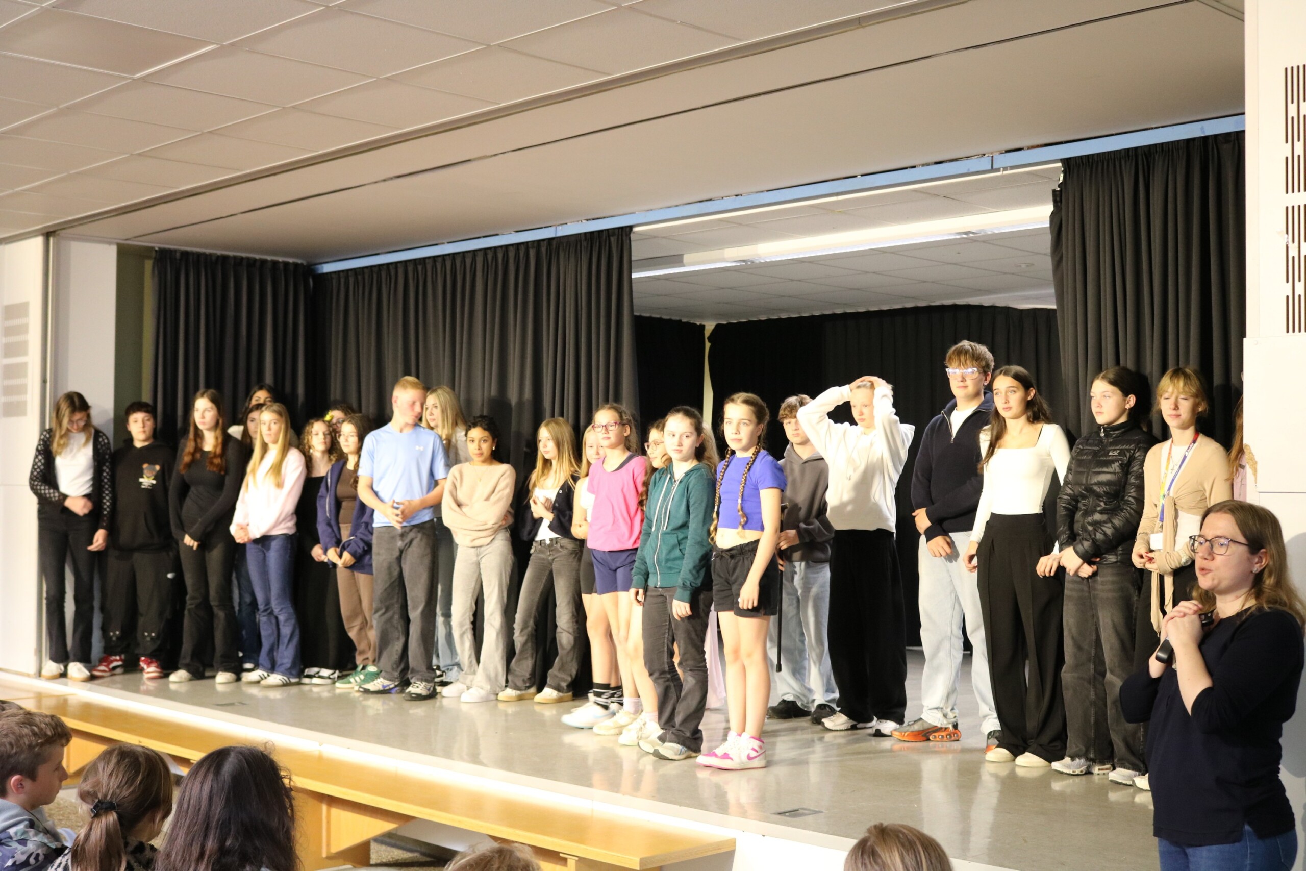 Alle Mitwirkenden der Open Stage – Schülerinnen und Schüler verschiedener Klassen sowie der DG-Kurs 9 – stehen gemeinsam auf der Bühne. Sie bilden eine lange Reihe und lassen sich vom Publikum feiern. Am rechten Bühnenrand ist eine Lehrkraft mit Mikrofon zu sehen, die die Veranstaltung moderiert.