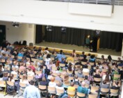 Anne Mauracher aus der Klasse 6d sitzt am Klavier und spielt die Stücke „Zerflossene Melodie“ und „Tanz der Intervalle“. Vor ihr füllt sich das Forum mit den 5. Klassen, die dicht gedrängt auf Stühlen sitzen und gespannt zuhören. Das Foto zeigt die gesamte Probebühne im architektonischen Rahmen des Forums.