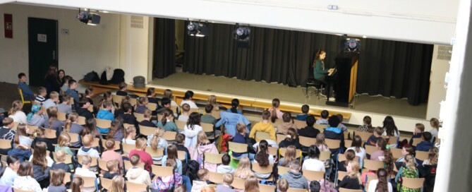 Anne Mauracher aus der Klasse 6d sitzt am Klavier und spielt die Stücke „Zerflossene Melodie“ und „Tanz der Intervalle“. Vor ihr füllt sich das Forum mit den 5. Klassen, die dicht gedrängt auf Stühlen sitzen und gespannt zuhören. Das Foto zeigt die gesamte Probebühne im architektonischen Rahmen des Forums.