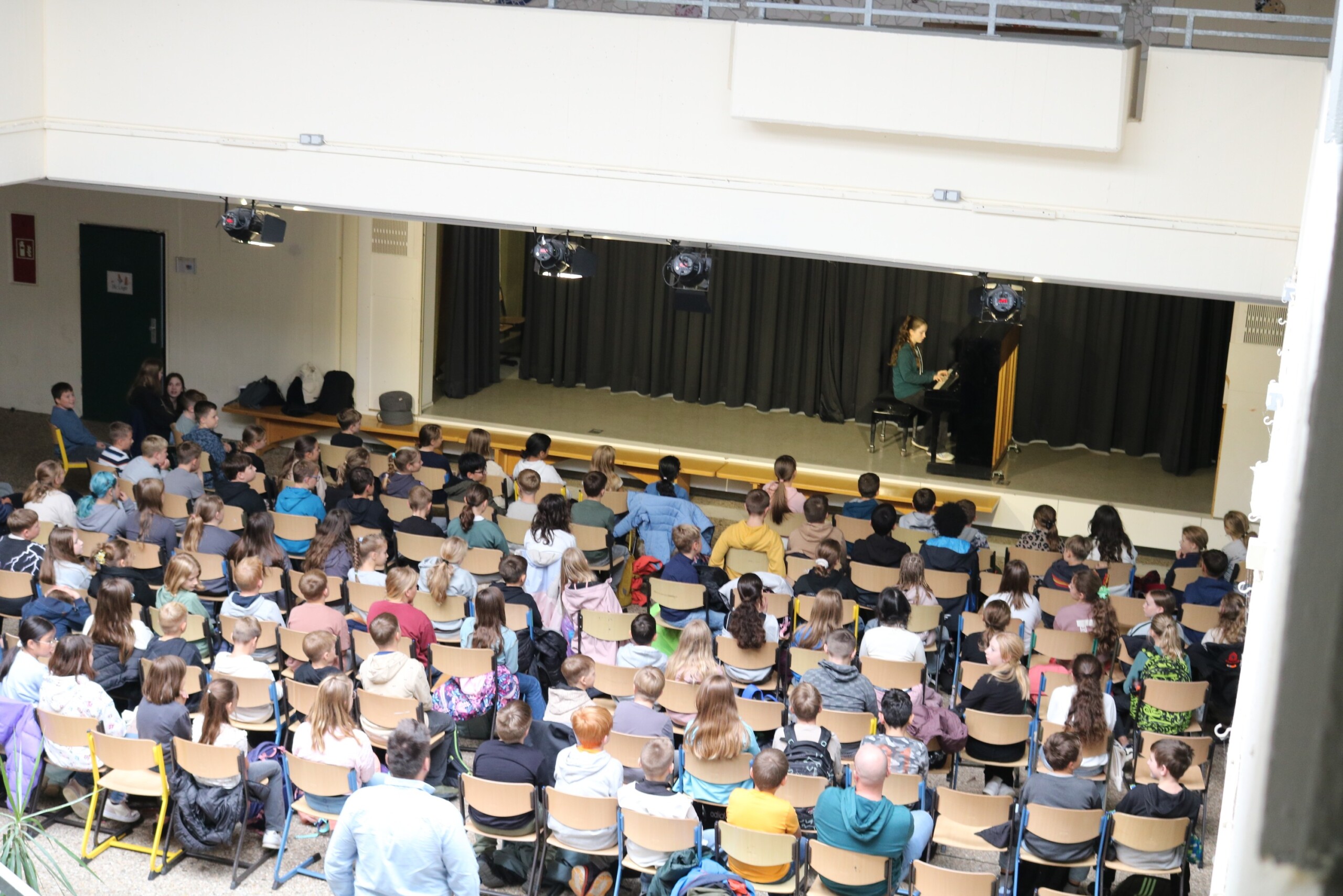 Anne Mauracher aus der Klasse 6d sitzt am Klavier und spielt die Stücke „Zerflossene Melodie“ und „Tanz der Intervalle“. Vor ihr füllt sich das Forum mit den 5. Klassen, die dicht gedrängt auf Stühlen sitzen und gespannt zuhören. Das Foto zeigt die gesamte Probebühne im architektonischen Rahmen des Forums.