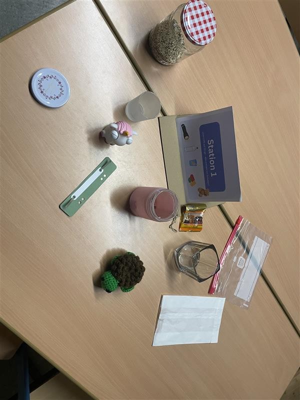 Gegenstände einer Lernstation liegen auf einem Tisch, darunter ein Glas, ein Jutebeutel, ein Smoothieglas, ein Schraubglas und ein Zettel mit „Station 1“.