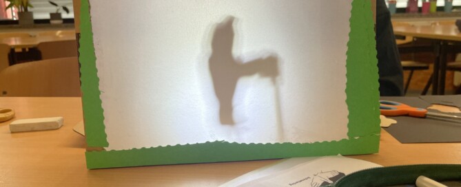 Ein selbstgebautes Schattentheater steht auf einem Tisch, dahinter ist die Silhouette einer Figur zu erkennen.