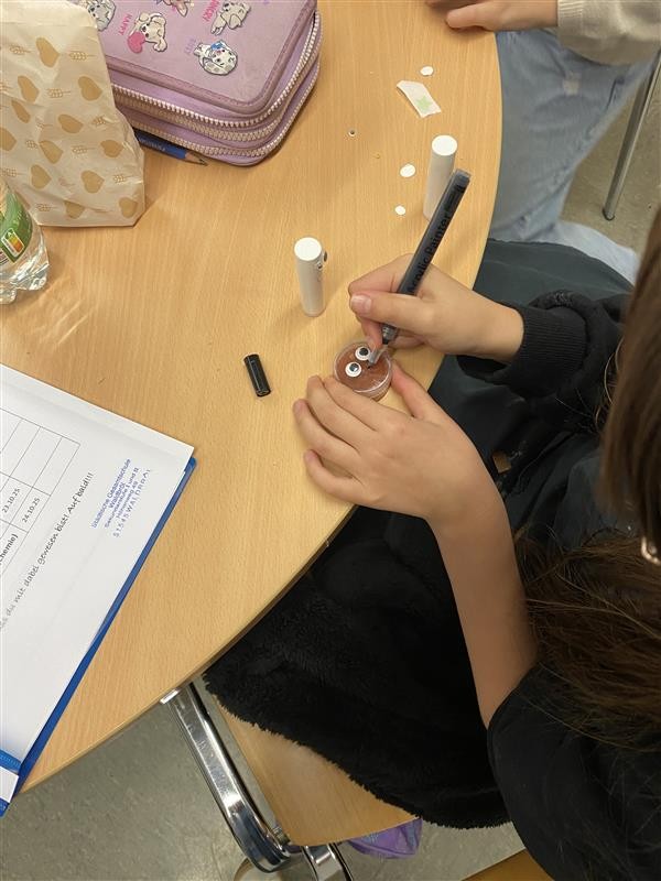 Ein Kind verziert einen kleinen Cremetiegel mit aufgekleben Wackelaugen und einem Stift.