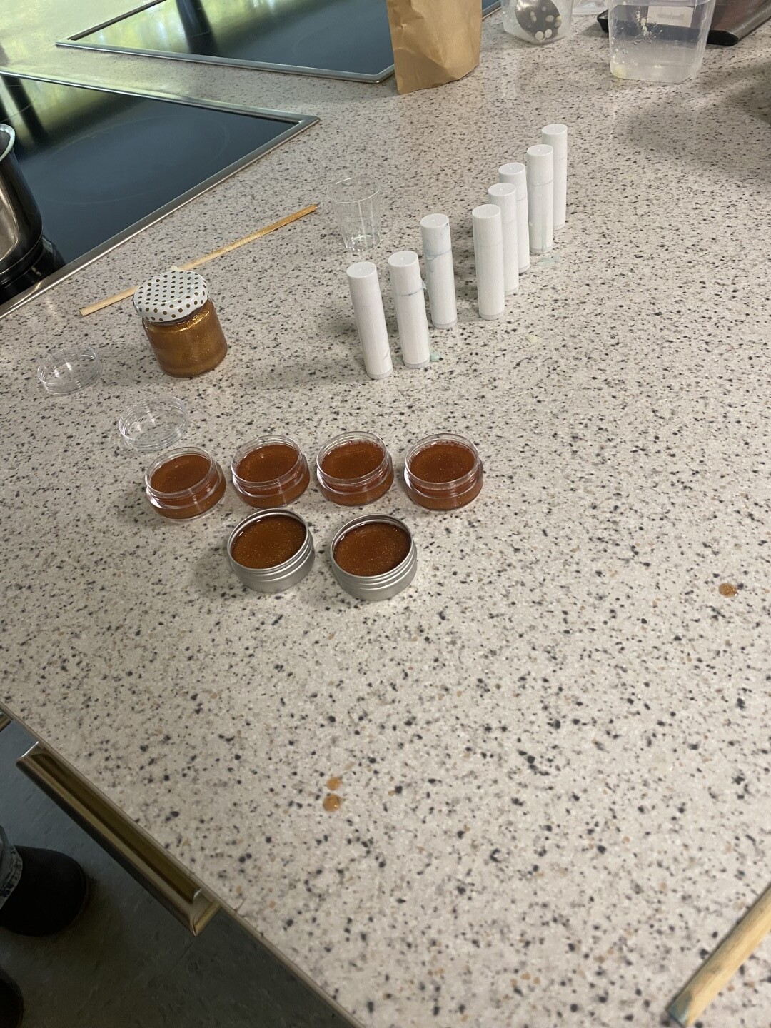 Mehrere geöffnete Kosmetiktiegel mit brauner Creme stehen auf einer Arbeitsfläche, daneben leere Lippenpflegestifthüllen.