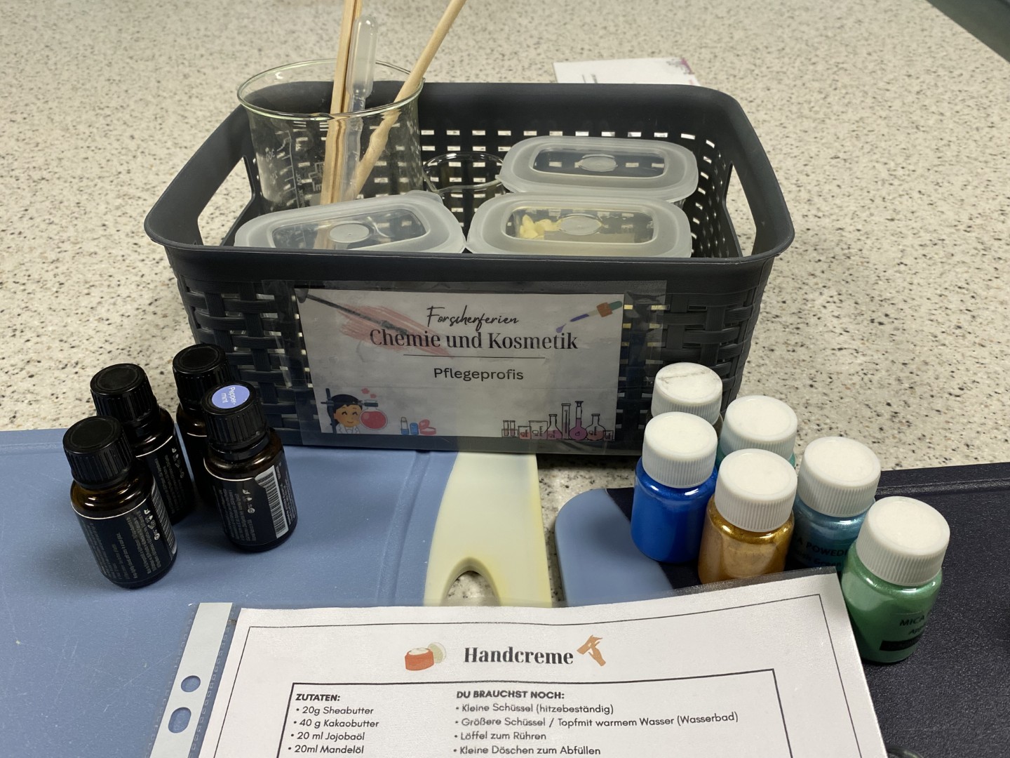 Materialien für den Workshop „Chemie und Kosmetik“, darunter Bechergläser, ätherische Öle, Farbpigmente und ein Rezept für Handcreme.