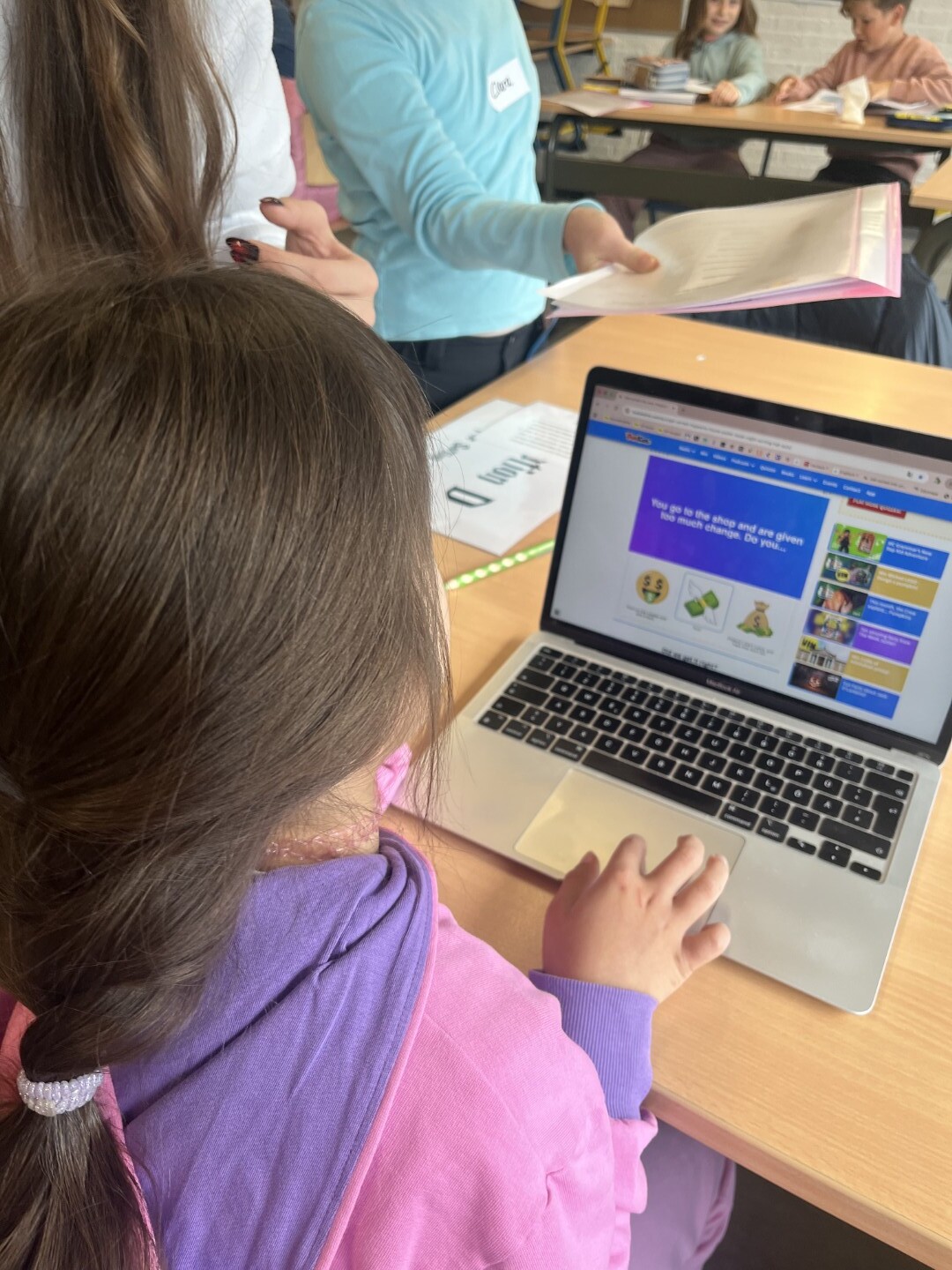 Ein Kind arbeitet an einem Laptop und löst eine digitale Aufgabe, daneben hält ein anderes Kind ein Heft.