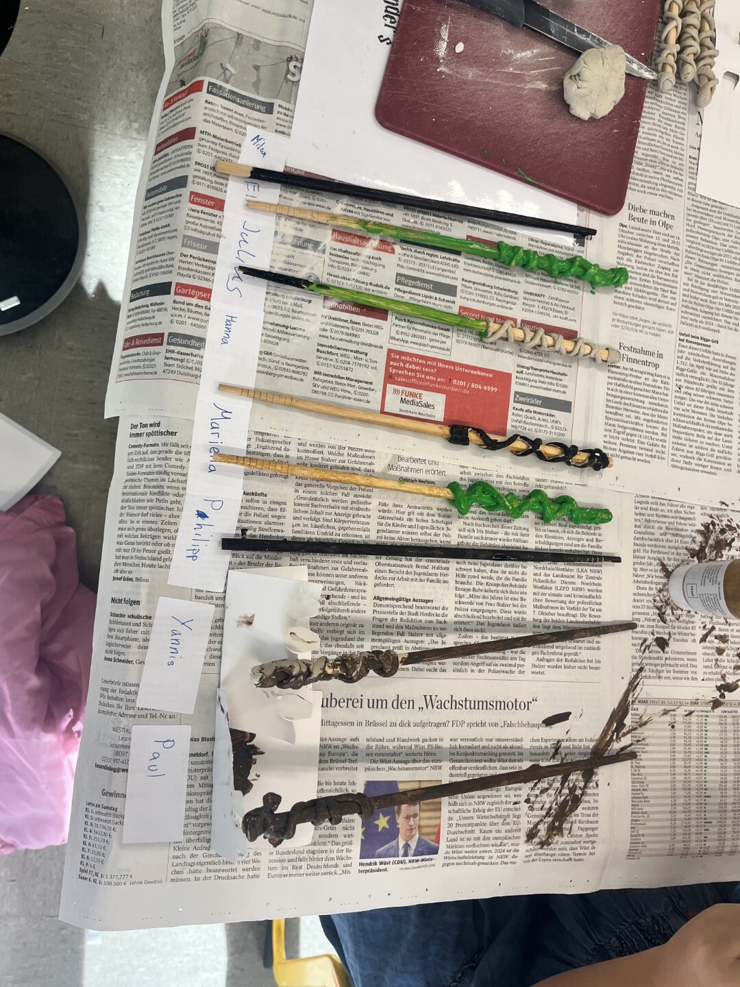 Eine Reihe selbstgebastelter Zauberstäbe liegt zum Trocknen auf Zeitungspapier, jeder Stab ist mit einem Namenszettel versehen.