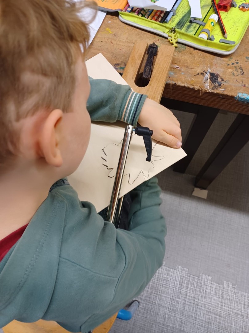 Ein Kind arbeitet konzentriert mit einer Laubsäge an einer Holzfigur, auf dem Tisch liegen bunte Stifte und Werkzeuge.