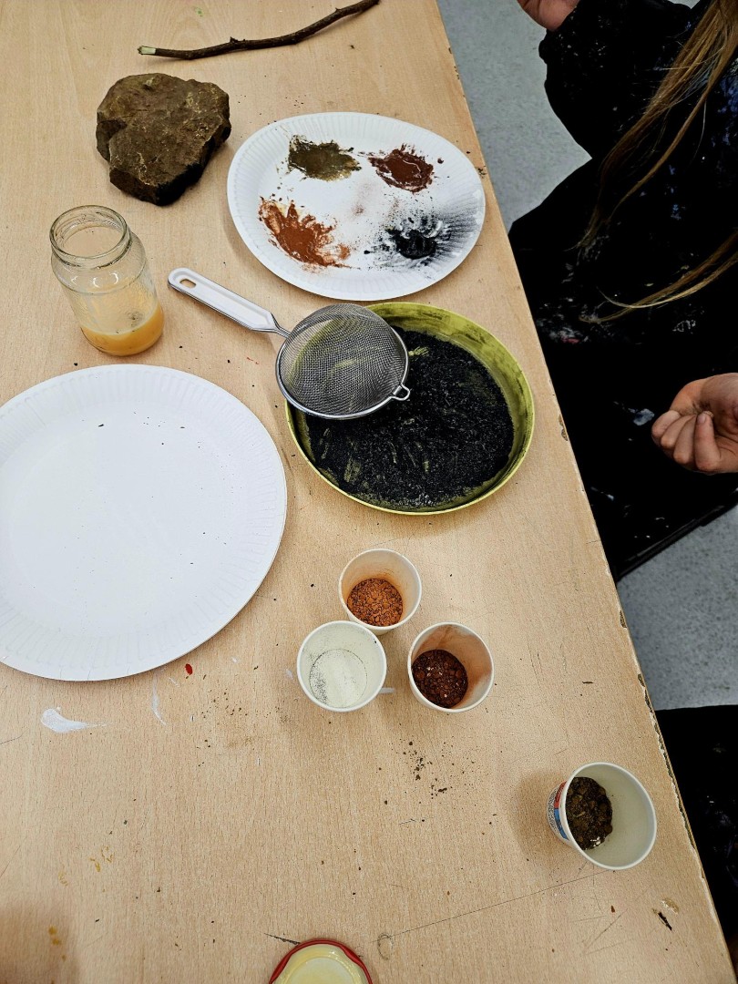 Naturpigmente in kleinen Bechern, Erdfarben auf Papieren sowie ein Sieb und Werkmaterialien liegen für das Herstellen von Höhlenfarben bereit.