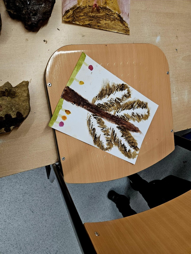 Ein Kinderbild mit einem Baum, gemalt in Naturfarben, liegt auf einem Stuhl neben weiteren Werkstücken.