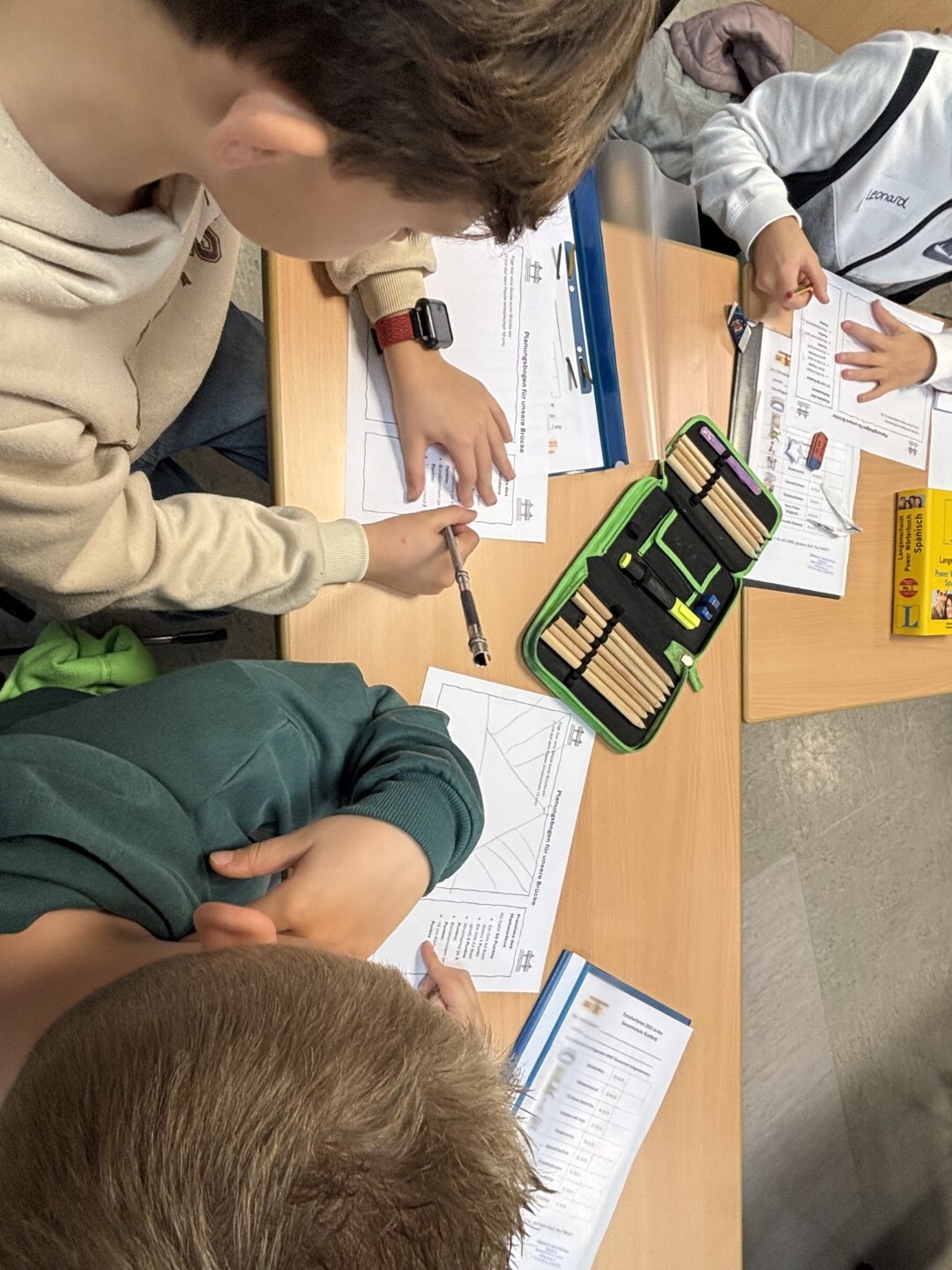 Drei Kinder sitzen an einem Tisch und planen auf Arbeitsblättern den Bau einer Papierbrücke.