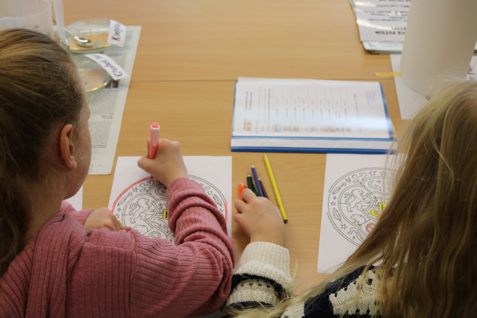 Zwei Kinder kolorieren ein Wappenmotiv auf einem Arbeitsblatt mit Buntstiften.