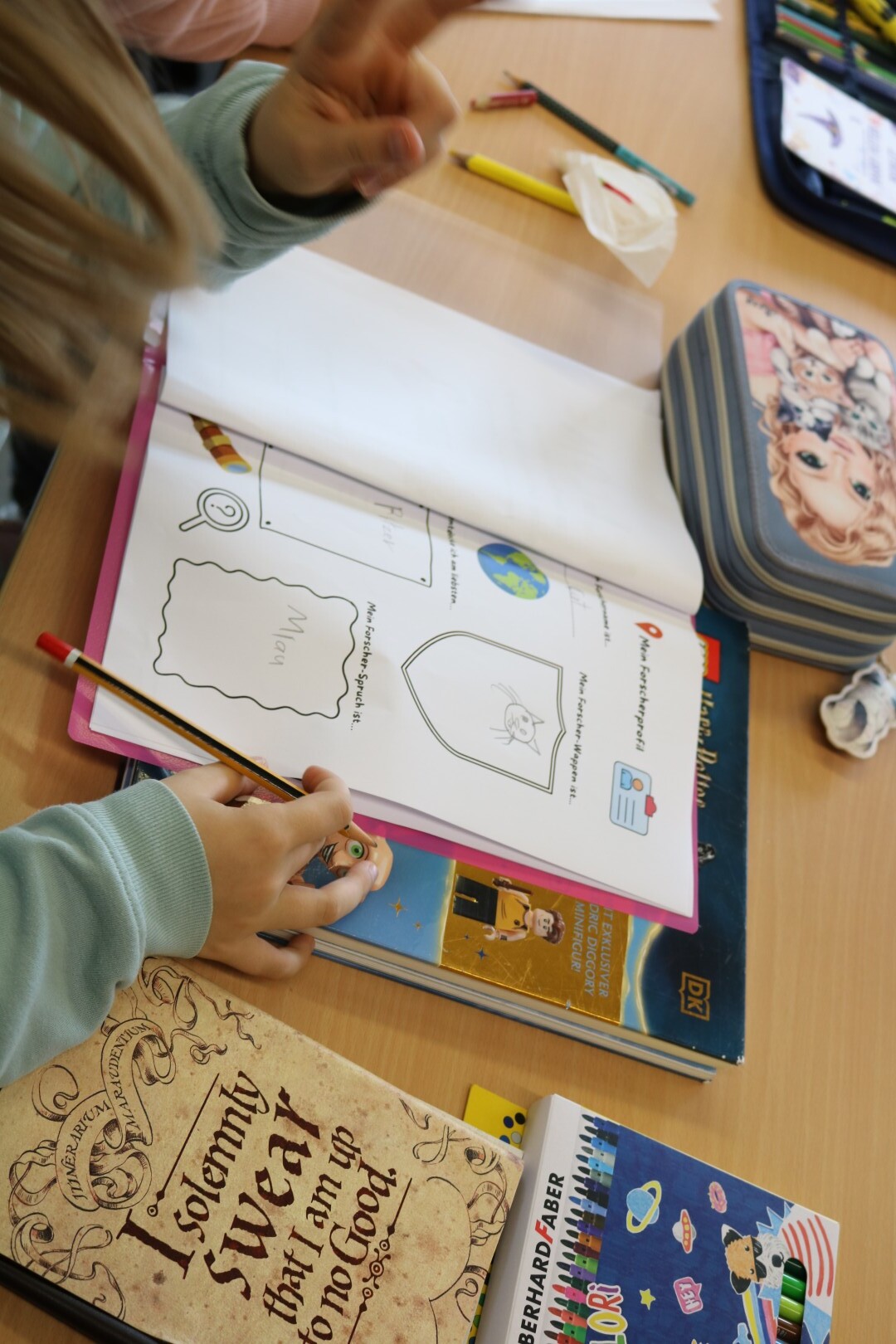 Ein Kind arbeitet in einem Heft mit dem Titel „Mein Forscherprofil“, daneben liegen Bücher und Stifte auf dem Tisch.