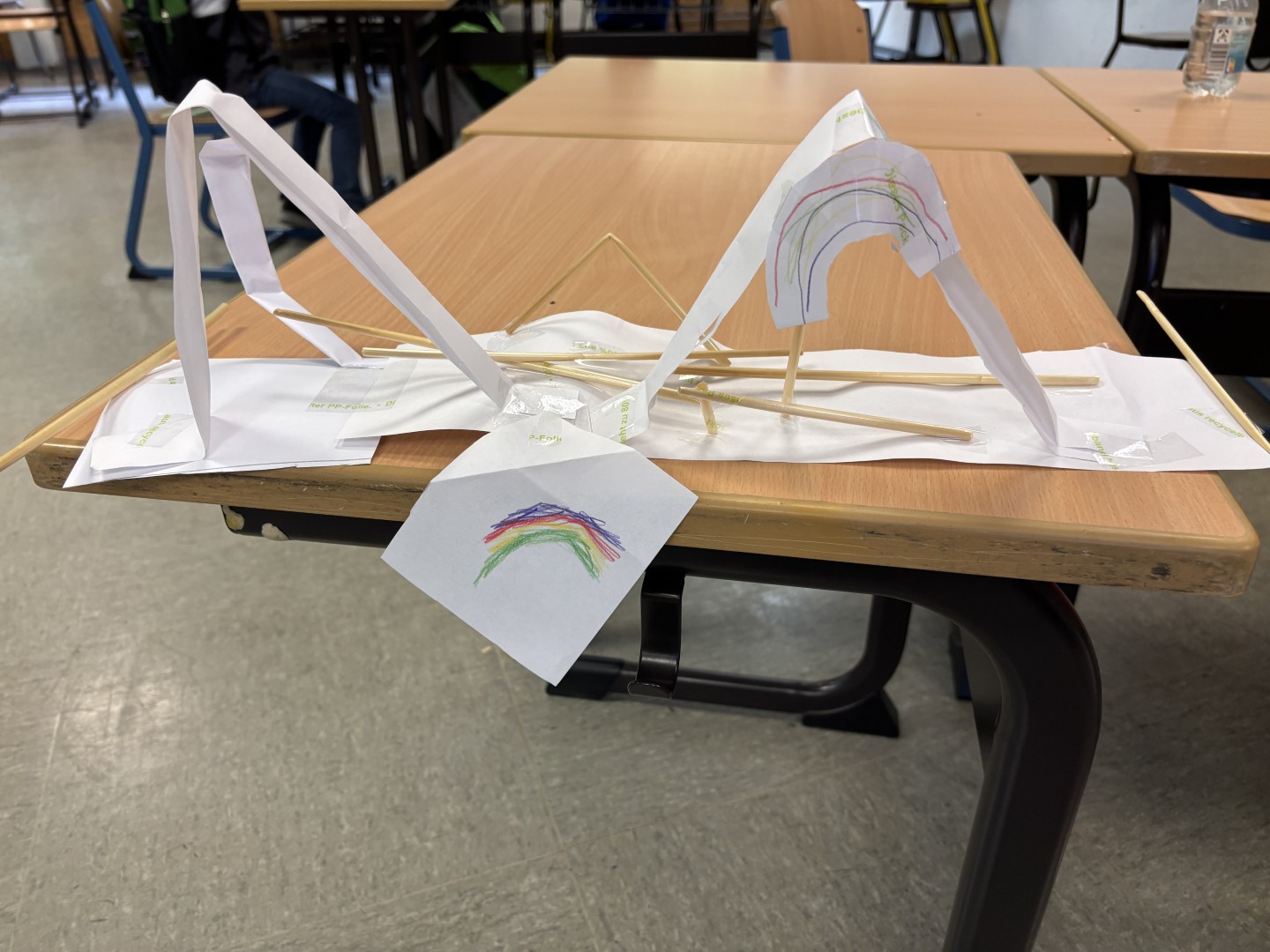 Eine bunte Papierbrücke mit Regenbogenzeichnung verbindet zwei Tische im Klassenraum.