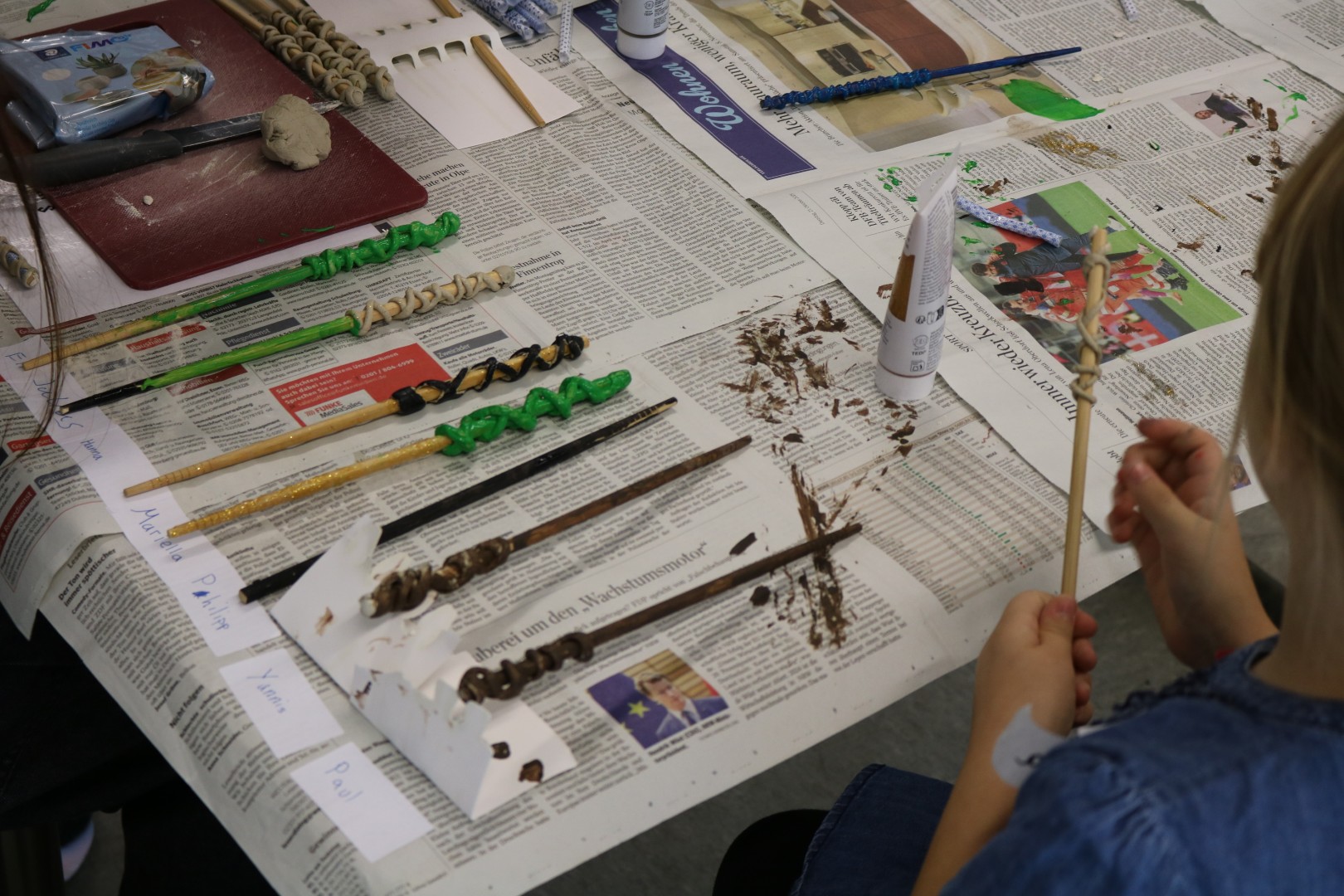 Gebastelte Zauberstäbe aus Holz und Knete liegen zum Trocknen auf Zeitungspapier, ein Kind bemalt einen Stab.