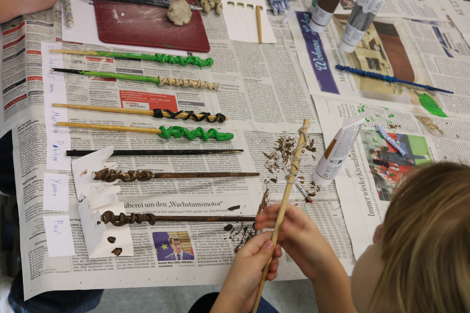 Weitere bemalte Zauberstäbe liegen auf Zeitungspapier, ein Kind dekoriert einen Stab mit modellierter Verzierung.