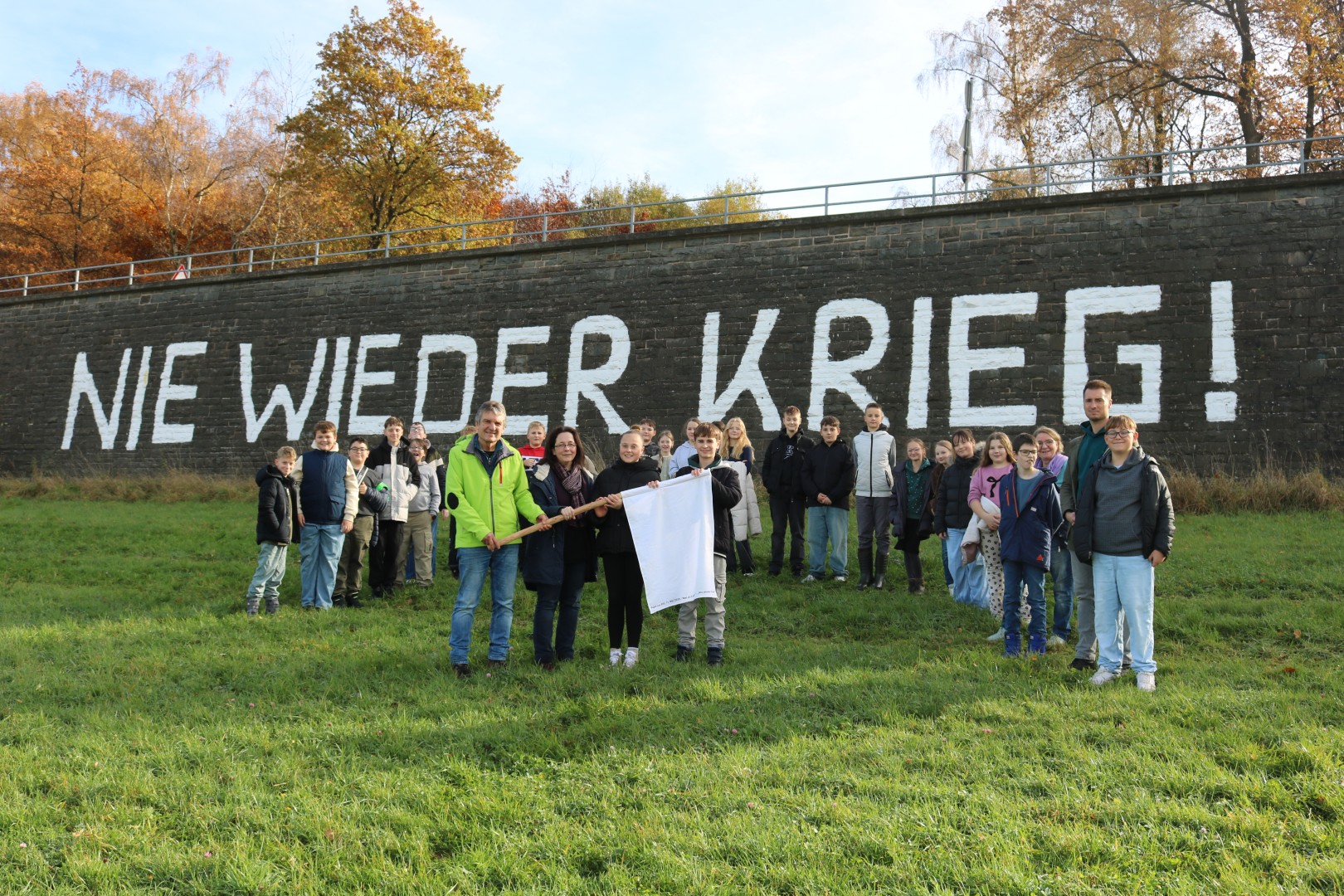 „Gruppenfoto an der Waldbröler Friedensmauer mit dem Schriftzug ‚NIE WIEDER KRIEG!‘. Vorne überreichen Johannes Heibel und zwei Schüler:innen die Weiße Friedensfahne. Hinter ihnen stehen die Schüler:innen der Klasse 6c.“