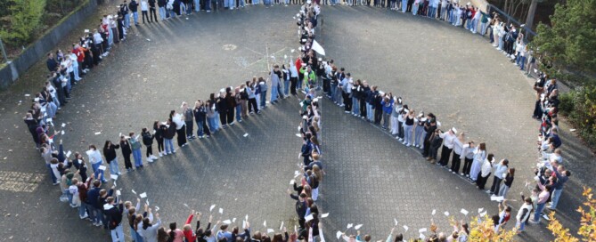 „Rund 300 Schüler:innen stehen auf dem Schulhof der Gesamtschule Waldbröl in einer großen Formation und bilden ein Peace-Zeichen. Viele halten kleine weiße Fahnen in die Höhe.“