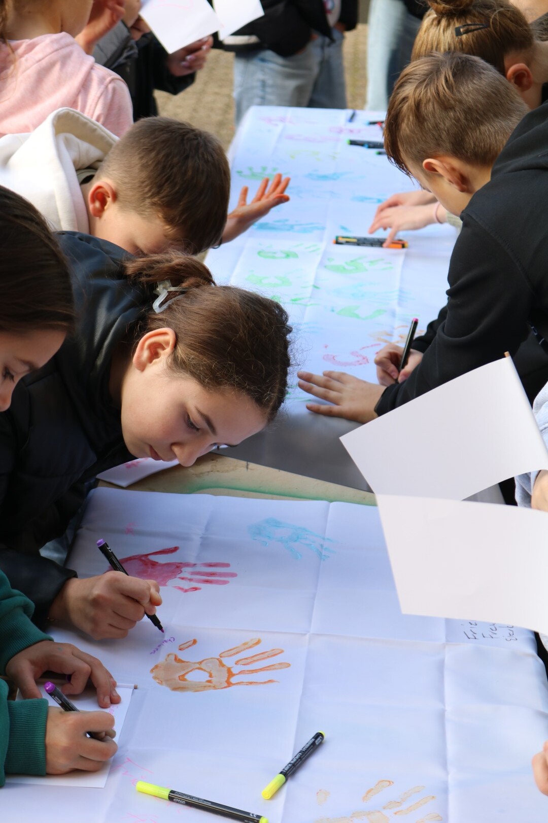 „Schüler:innen beugen sich über ein langes weißes Tuch und gestalten es mit bunten Handabdrücken und kurzen Friedensbotschaften.“