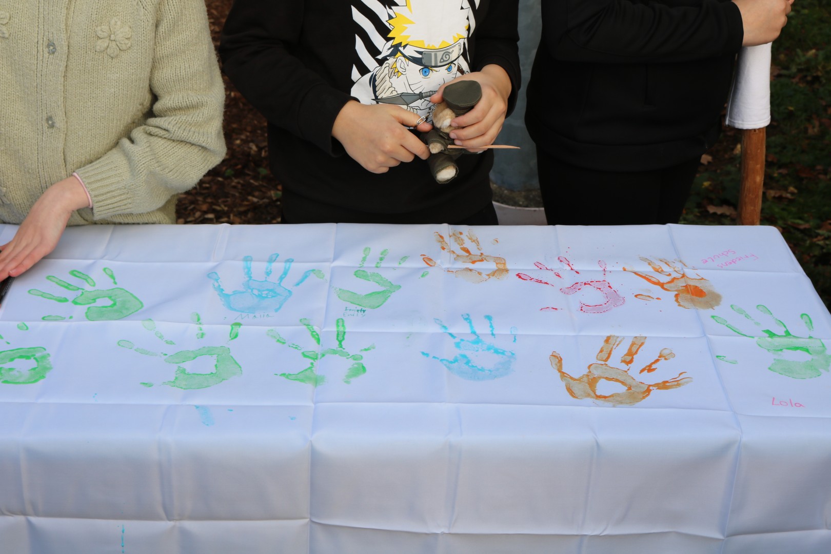 „Ein Abschnitt des weißen Friedensbands mit bunten Handabdrücken von Schüler:innen. Im Bild sind die Hände und Oberkörper mehrerer Kinder zu sehen, die das gestaltete Tuch präsentieren.“