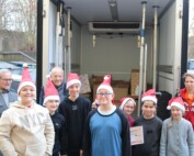 Eine Gruppe von neun Schüler:innen mit Weihnachtsmützen steht vor einem geöffneten Lieferwagen der Tafel Oberberg Süd. Hinter ihnen sind mehrere Kartons sichtbar. Zwei erwachsene Mitarbeitende der Tafel sowie eine erwachsene Begleitperson stehen am Rand. Ein Kind hält eine Urkunde in der Hand.