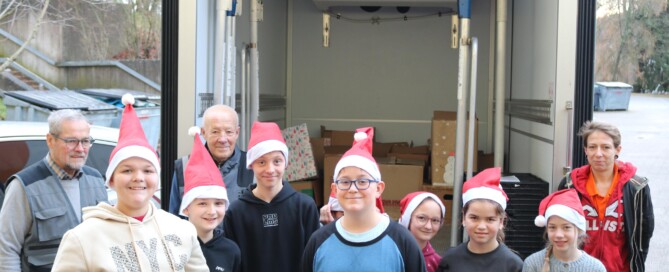 Eine Gruppe von neun Schüler:innen mit Weihnachtsmützen steht vor einem geöffneten Lieferwagen der Tafel Oberberg Süd. Hinter ihnen sind mehrere Kartons sichtbar. Zwei erwachsene Mitarbeitende der Tafel sowie eine erwachsene Begleitperson stehen am Rand. Ein Kind hält eine Urkunde in der Hand.