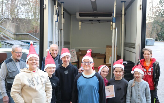 Eine Gruppe von neun Schüler:innen mit Weihnachtsmützen steht vor einem geöffneten Lieferwagen der Tafel Oberberg Süd. Hinter ihnen sind mehrere Kartons sichtbar. Zwei erwachsene Mitarbeitende der Tafel sowie eine erwachsene Begleitperson stehen am Rand. Ein Kind hält eine Urkunde in der Hand.