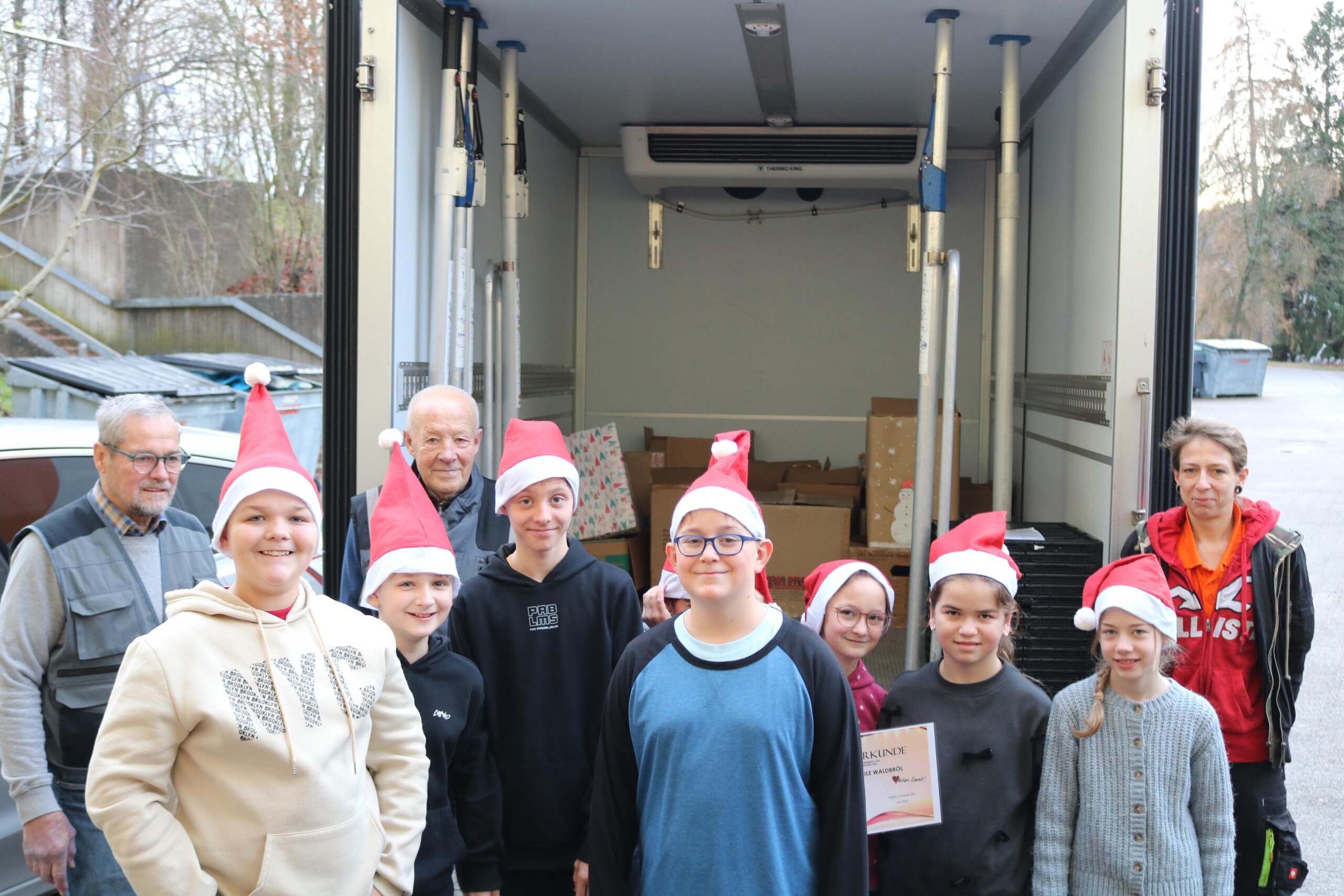 Eine Gruppe von neun Schüler:innen mit Weihnachtsmützen steht vor einem geöffneten Lieferwagen der Tafel Oberberg Süd. Hinter ihnen sind mehrere Kartons sichtbar. Zwei erwachsene Mitarbeitende der Tafel sowie eine erwachsene Begleitperson stehen am Rand. Ein Kind hält eine Urkunde in der Hand.