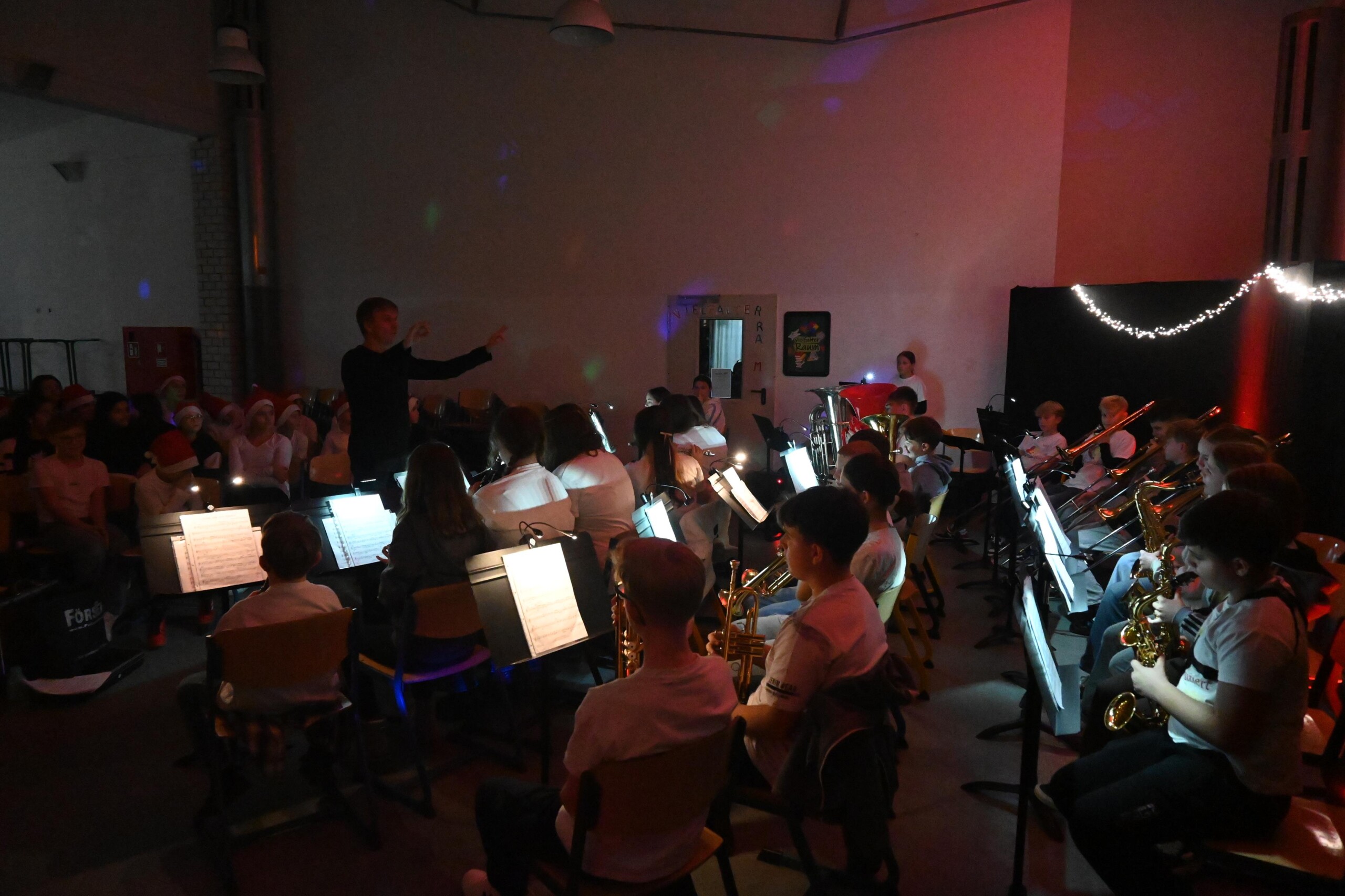 Bläserklasse der Gesamtschule sitzt halbrund auf der Bühne, dirigiert von einem Lehrer, mit Notenständern und Blasinstrumenten, stimmungsvolle Beleuchtung während der Veranstaltung „Lichter im Advent“.