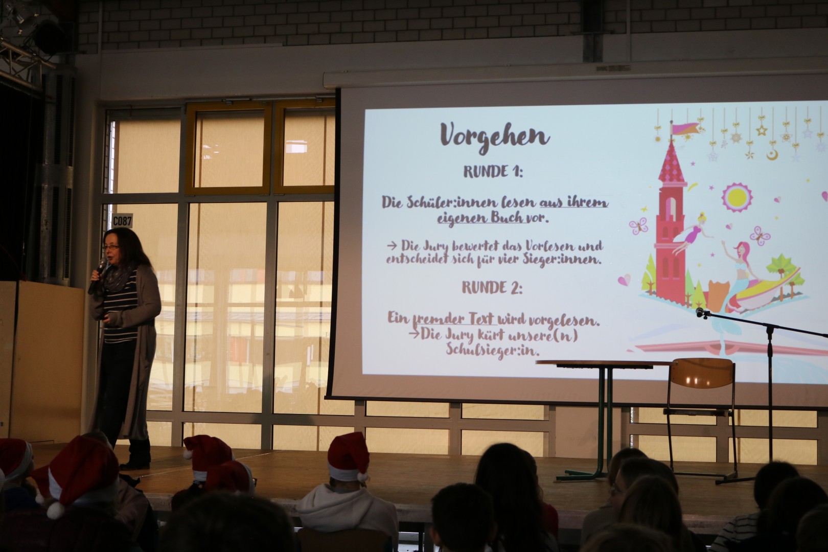 Moderatorin steht auf der Bühne und erklärt den Ablauf des Vorlesewettbewerbs vor einer projizierten Folie mit dem Titel „Vorgehen“.