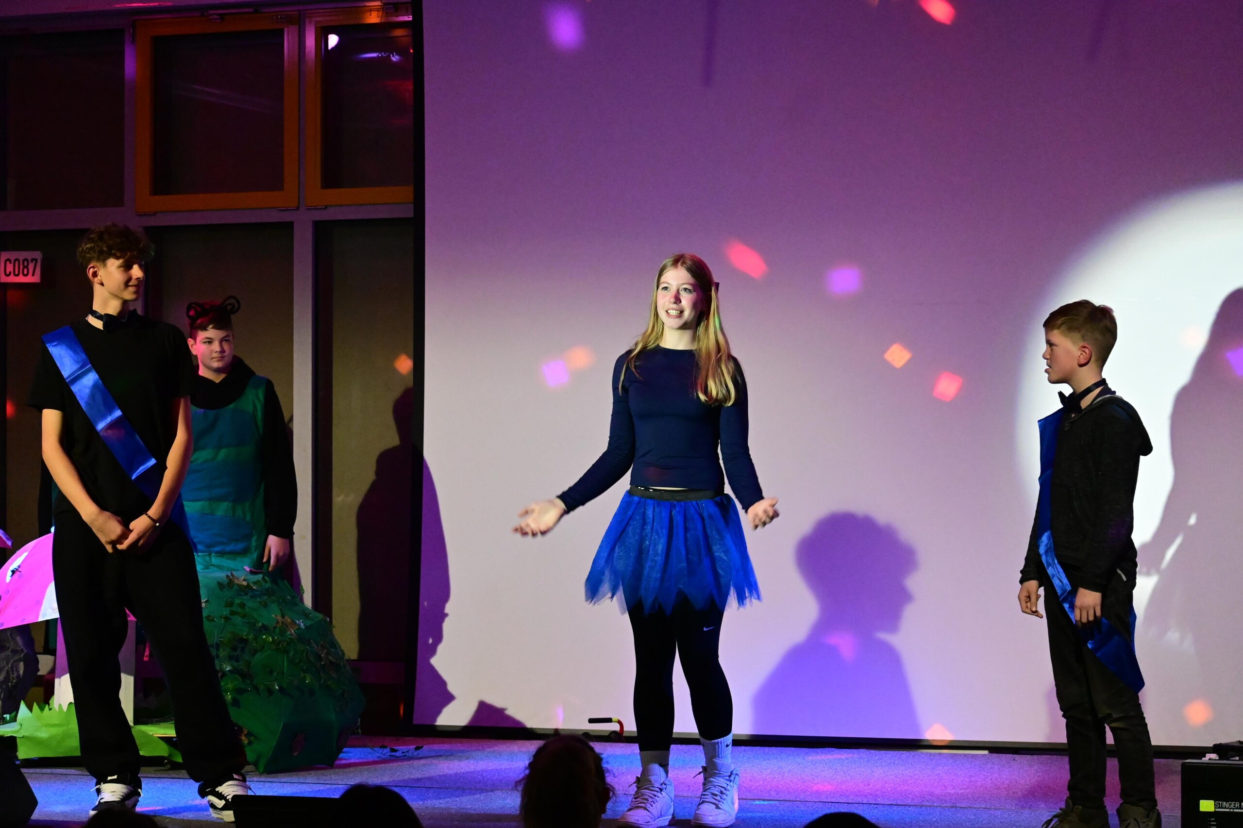 Szenische Darstellung mit drei Schüler:innen auf der Bühne, eine Schülerin steht mittig und spricht, zwei weitere stehen seitlich, Bühnenlicht erzeugt farbige Akzente.