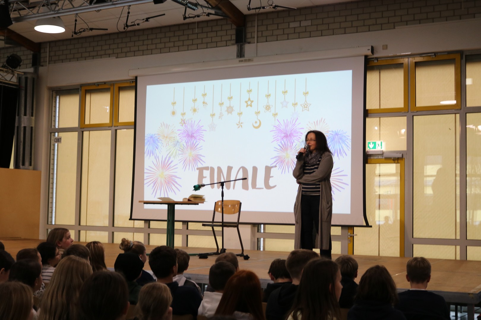 Moderatorin steht auf der Bühne vor der Folie „Finale“ mit Feuerwerksgrafiken; das Publikum schaut aufmerksam zu.