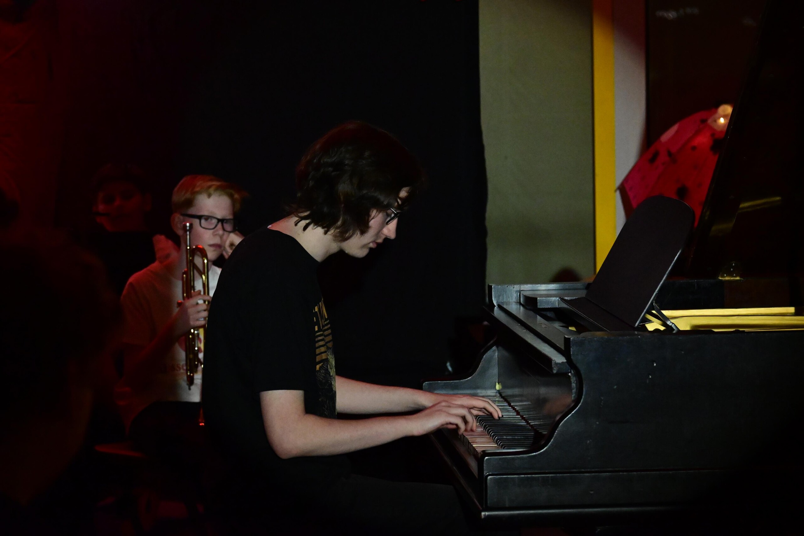 Schüler spielt Klavier auf der Bühne, im Hintergrund steht ein weiterer Schüler mit Trompete, dunkle Bühnenbeleuchtung.