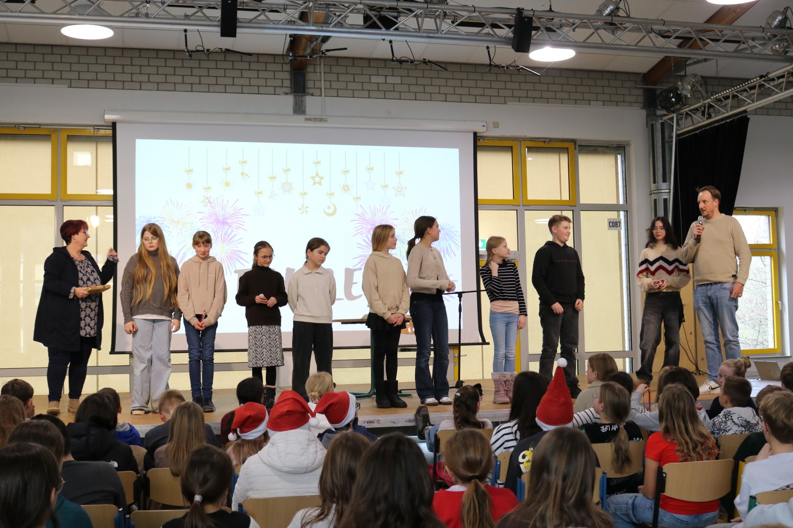 Acht Schüler:innen stehen nebeneinander auf der Bühne vor einer Leinwand mit der „Finale“-Folie; eine Moderatorin und ein Jurymitglied sprechen ins Mikrofon, das Publikum sitzt aufmerksam davor in der Mensa.