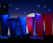 Mehrere Schüler:innen stehen nebeneinander auf der Bühne, vollständig in farbige Stoffe gehüllt, blaues und rotes Licht erzeugt eine abstrakte Bühnenwirkung.