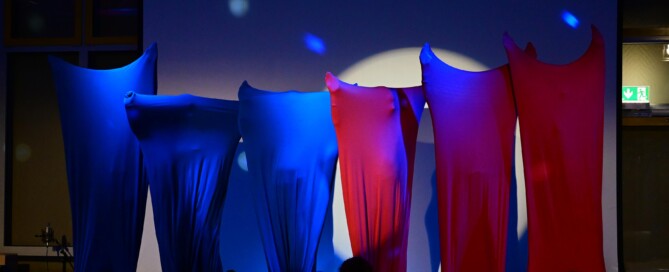 Mehrere Schüler:innen stehen nebeneinander auf der Bühne, vollständig in farbige Stoffe gehüllt, blaues und rotes Licht erzeugt eine abstrakte Bühnenwirkung.