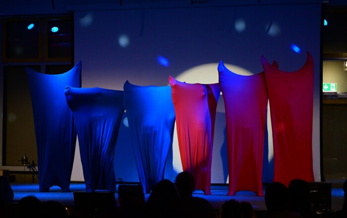 Mehrere Schüler:innen stehen nebeneinander auf der Bühne, vollständig in farbige Stoffe gehüllt, blaues und rotes Licht erzeugt eine abstrakte Bühnenwirkung.