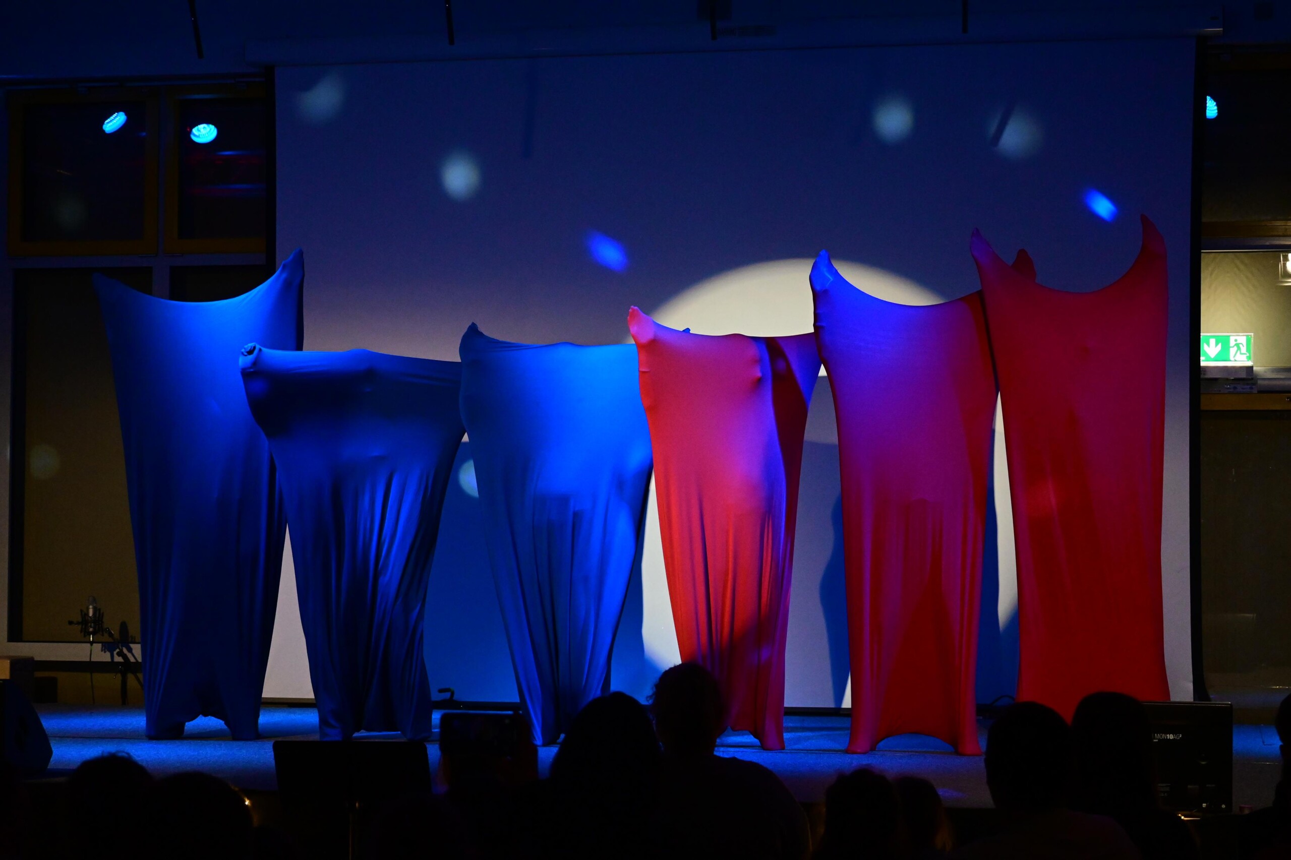 Mehrere Schüler:innen stehen nebeneinander auf der Bühne, vollständig in farbige Stoffe gehüllt, blaues und rotes Licht erzeugt eine abstrakte Bühnenwirkung.