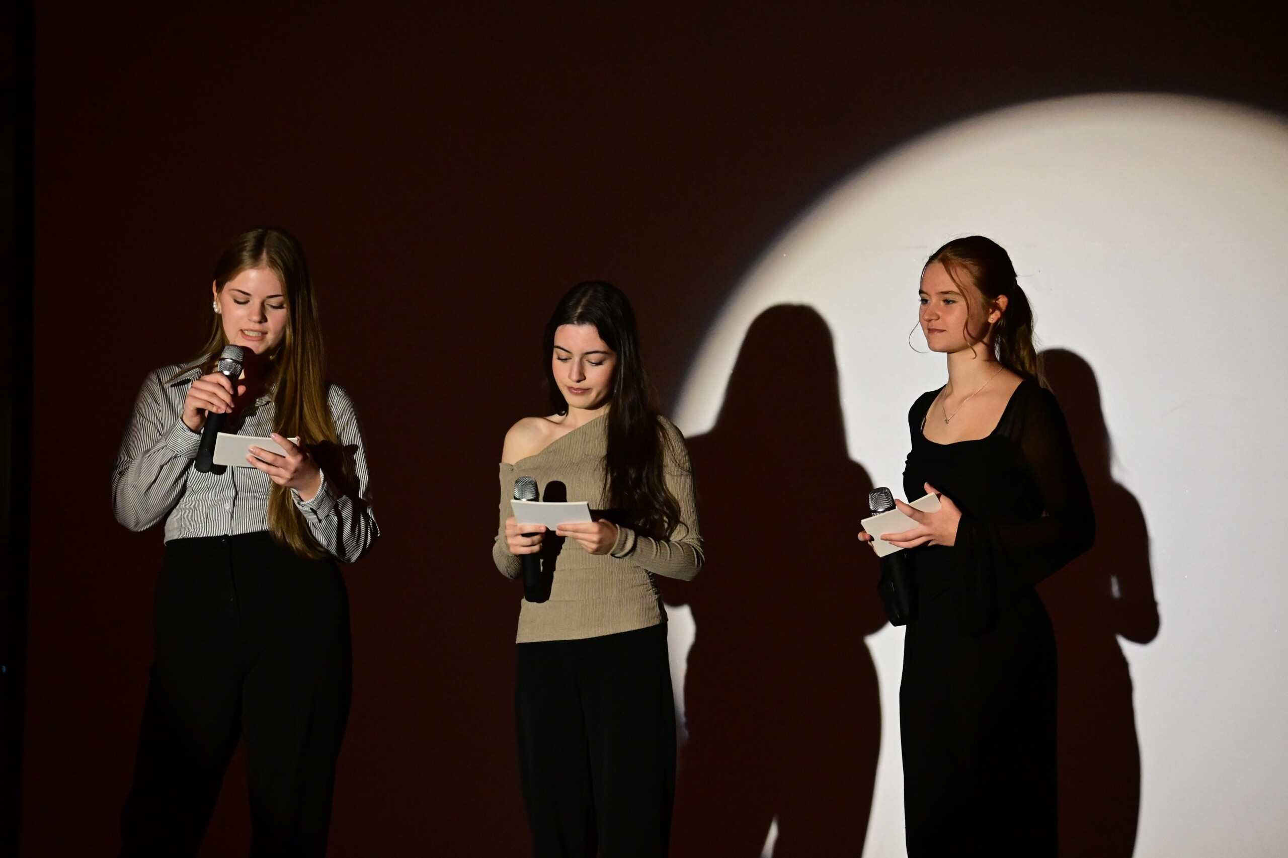 Drei Schülerinnen moderieren die Veranstaltung auf der Bühne, jede hält ein Mikrofon und eine Moderationskarte, heller Spot im Hintergrund.