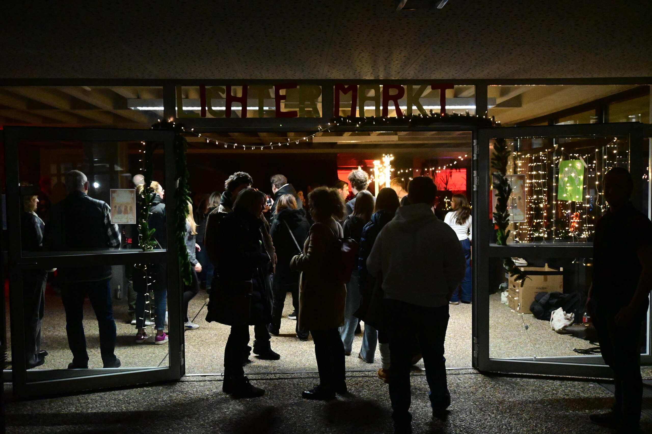 Blick auf den Eingangsbereich der Schule mit geöffneten Türen, viele Besucher:innen stehen und unterhalten sich vor dem beleuchteten Lichtermarkt.