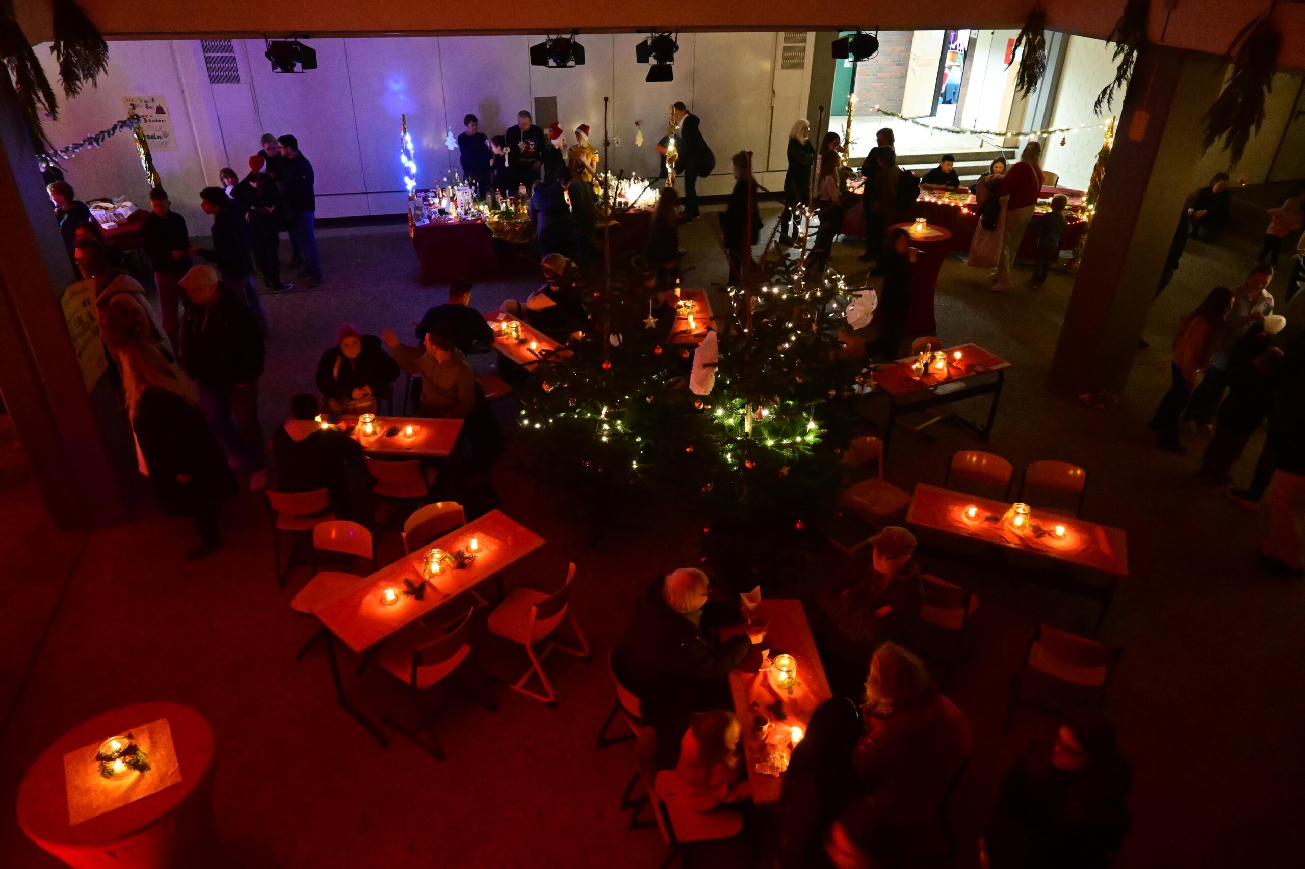 Blick von oben auf den Lichtermarkt im Schulgebäude, Tische mit Kerzen und Dekoration, Besucher:innen sitzen und stehen im Raum.