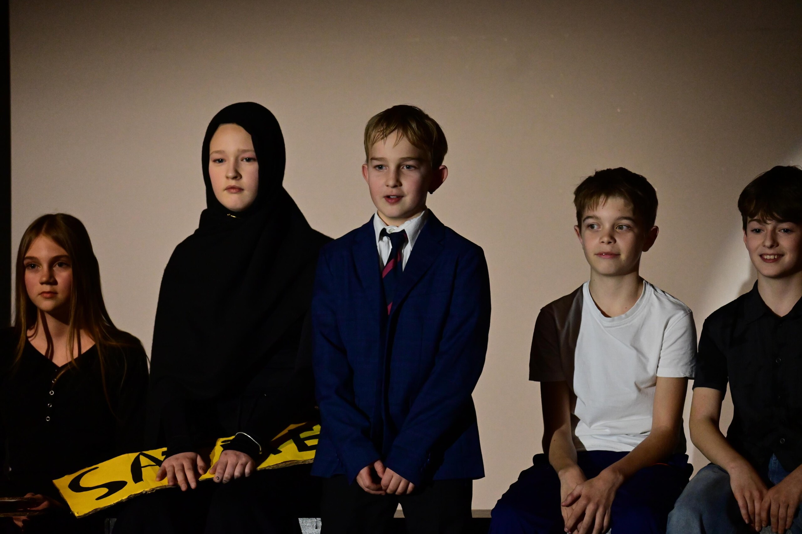 Mehrere Schüler:innen sitzen nebeneinander auf der Bühne und stellen eine Szene dar, unterschiedliche Kostüme deuten verschiedene Rollen an.